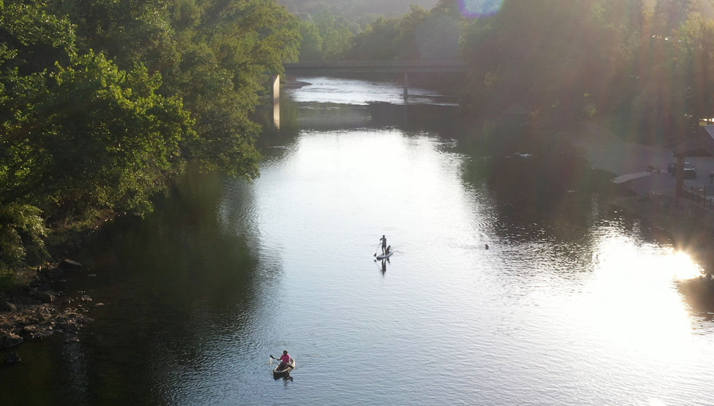 Paddlers on the Greenbrier River near Lewisburg, West Virginia, gliding through calm, reflective water at sunset beneath a riverside bridge and tree-lined banks.