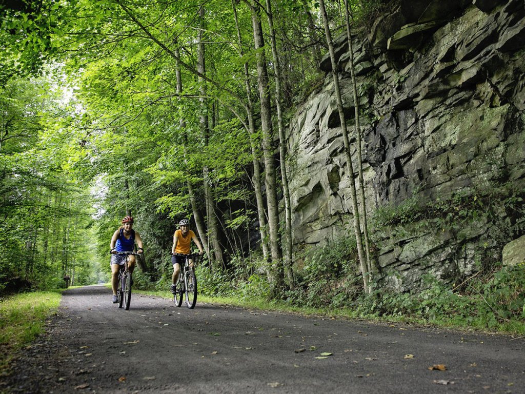 Two cyclists biking on the scenic Greenbrier River Trail past a rocky forested outcrop in Greenbrier Valley, West Virginia.