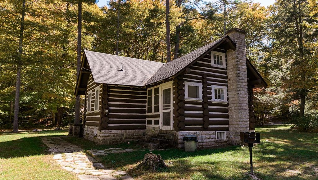 A rustic log cabin at Greenbrier State Forest, surrounded by tall trees and vibrant autumn foliage, featuring a stone chimney and a stone pathway.