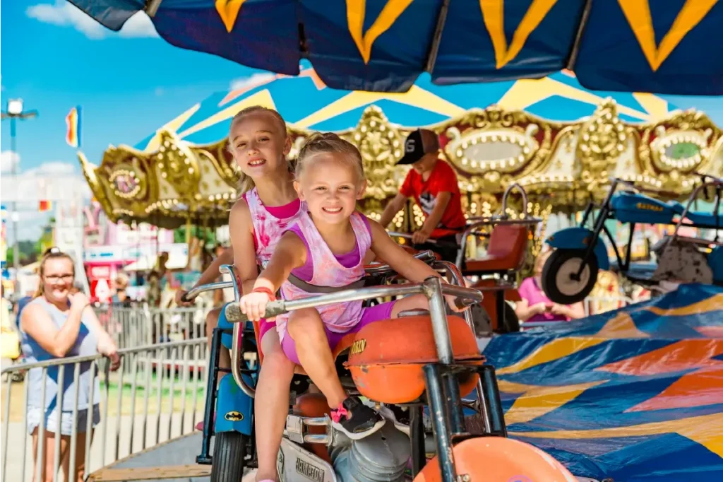 Two young girls joyfully ride a colorful carnival ride at the State Fair of West Virginia. They are surrounded by vibrant tents and a carousel in the background.