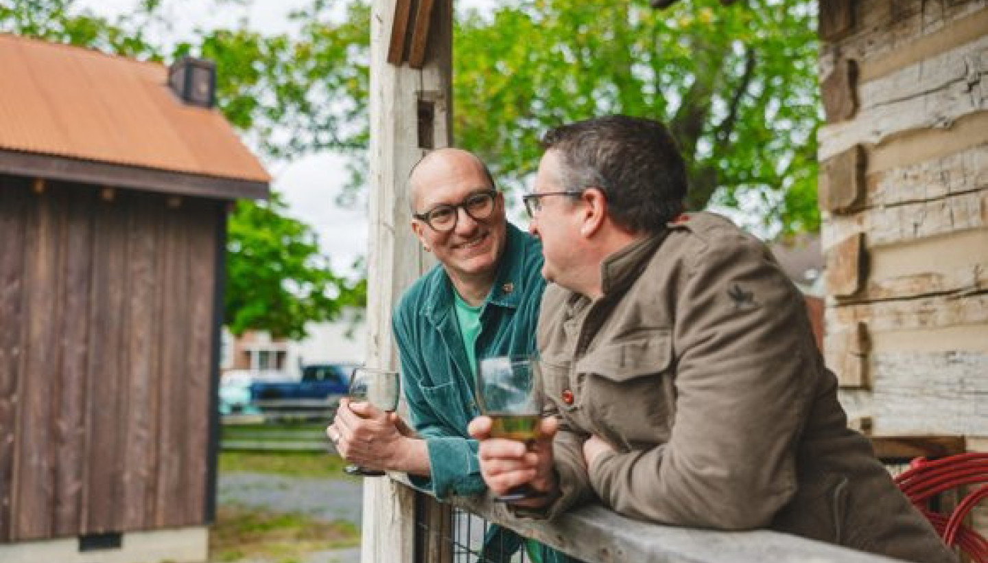 Couple enjoy drinks at Barnwood Village in White Sulphur Springs while leaning on a wooden railing.